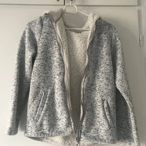 Grå fleecejacka med huva - Mysig grå fleecejacka med huva och dragkedja. Insidan är fodrad med mjuk teddyfleece för extra värme. Perfekt för kyliga dagar.