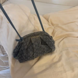 Blå axelväska i jeansmaterial - Säljer en snygg blå axelväska i jeansmaterial med en unik design. Väskan har en enkel axelrem och en intressant struktur som ger den en cool look. Perfekt för att ge din outfit en avslappnad touch.