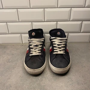 Svarta sneakers från Moncler - Snygga svarta sneakers från Moncler med vita snören och en röd detalj på sidan. Skorna har en klassisk design med en låg profil och är perfekta för en stilren look. Priset är inte hugget i sten!