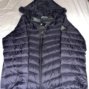 Svart dunjacka från Stone Island - Snygg svart dunjacka från Stone Island med huva och dragkedja. Jackan har ett quiltat mönster och en ikonisk logga på ärmen. Perfekt för kyliga dagar när du vill hålla stilen.