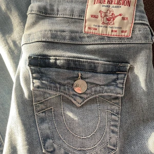 True Religion Joey Low Rise Flare Jeans - Trendiga True Religion jeans i en ljusblå tvätt med låg midja och flare-stil. De är i storlek 27 och i nyskick. Storleken är slutsålt på hemsidan. Nypris 1300kr