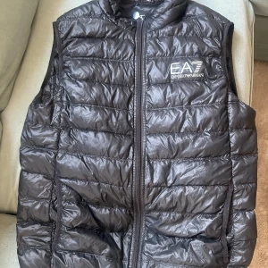 Svart dunväst från Emporio Armani - Snygg svart dunväst från Emporio Armani med dragkedja framtill. Perfekt för lager på lager-stil och håller dig varm under kyliga dagar. Västen har en stilren design med märkets logga på bröstet.