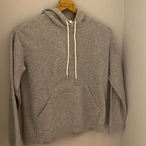Grå hoodie från H&M - Säljer en klassisk grå hoodie från H&M Basics. Den har en bekväm passform med långa ärmar och magficka. Perfekt för en avslappnad stil. Huvan har vita dragsnören för justering.