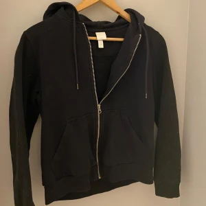 Svart ziphoodie från H&M - Säljer en svart ziphoodie från H&M med dragkedja och huva. Tröjan har långa ärmar och en praktisk ficka framtill. Perfekt för en avslappnad stil.