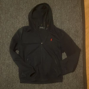 Mörkblå  jacka från Ralph Lauren - Säljer en stilren mörkblå jacka från Ralph Lauren med röd logga på bröstet. Den har dragkedja framtill och en praktisk huva med snörning. Perfekt för en casual look. Storlek på jackan är M men skulle säga att den passar bättre s liten i storlek alltså.