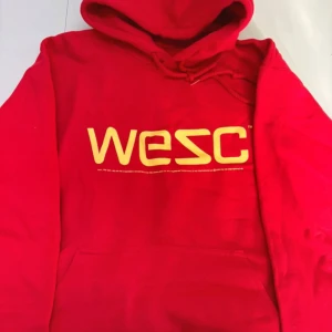 Röd hoodie från WESC - Säljer en cool röd hoodie från WESC med gult tryck på bröstet. Den har en klassisk känguruficka och justerbar huva med snören. Perfekt för en avslappnad stil.