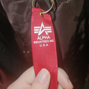 Brun bomberjacka från Alpha Industries - Säljer en snygg brun bomberjacka från Alpha Industries. Jackan har en dragkedja i guld och ribbade muddar vid ärmslut och nederkant. Den har en klassisk bomberstil med fickor och en röd tagg på ärmen. Perfekt för en cool och avslappnad look.