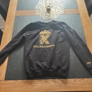 Svart tröja från Dolce & Gabbana - Säljer en svart tröja från Dolce & Gabbana med ett stort broderat 'K' och krona i guld på framsidan. Tröjan har långa ärmar och D&G-broderi på ärmen. Perfekt för dig som vill ha något stilrent och lyxigt. Priset är inte hugget i sten så fråga på