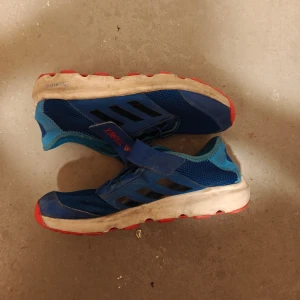 Blå sneakers från Adidas - Säljer ett par blå sneakers från Adidas med svarta detaljer och en vit sula. Skorna har en praktisk kardborreknäppning och är perfekta för vardagsbruk. De har en sportig design med en bekväm passform.