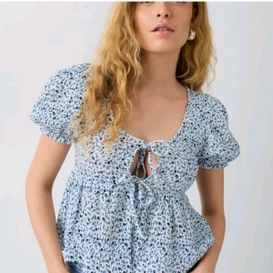 Blommig blus med knytning - Charmig blommig blus i blått och vitt med korta puffärmar och knytning framtill. Perfekt för en somrig look med sitt luftiga och lätta material. Inga defekter