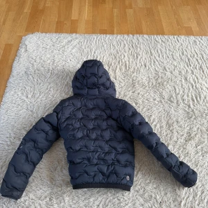 Marin blå dunjacka från Colmar - Säljer en snygg svart dunjacka från Colmar med huva och dragkedja. Jackan har en quiltad design och ribbade muddar vid ärmslut och nederkant. Perfekt för kyliga dagar!