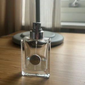 Versace Pour homme - Säljer en stilren och elegant parfymflaska i glas med en fyrkantig form. Flaskan har en silverfärgad spraypump och en rund silverdetalj på framsidan. Perfekt för den som gillar minimalistisk design.