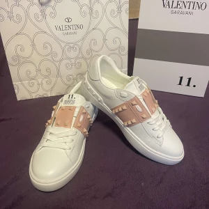 Valentino Garavani Rockstud sneakers - Snygga vita Valentino Garavani Rockstud sneakers med rosa detaljer och ikoniska nitar. Skorna har en stilren design med snörning och en bekväm passform. Perfekta för att ge din outfit en lyxig touch.
