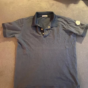 Mörkblå pikétröja från Moncler - Säljer en stilren mörkblå pikétröja från Moncler med klassisk krage och knappar framtill. Tröjan har korta ärmar och ett märke på ärmen. Står även Moncler på kragen. 100% äkta köpt på monclers hemsida. Tröjan finns inte längre det var en limited edition. Nypris 2050