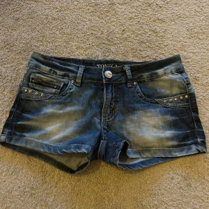 Blå jeansshorts från Toxik3 - Snygga blå jeansshorts från Toxik3 med slitna detaljer och nitar vid fickorna. De har en knapp och dragkedja framtill samt klassiska femficksdesign. Perfekta för en avslappnad stil.