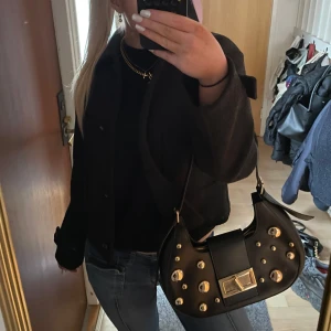 Svart axelväska med gulddetaljer - Snygg svart axelväska i skinn med coola guldfärgade nitar och ett stilrent spänne framtill. Väskan har en praktisk dragkedja och justerbar axelrem. 