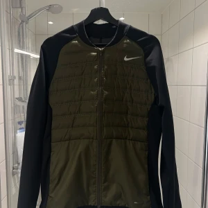 Olivgrön jacka från Nike - Snygg olivgrön jacka från Nike med svarta ärmar och dragkedja framtill. Jackan har en quiltad design på bröstet och ryggen, vilket ger en sportig look. Perfekt för höst och vår.