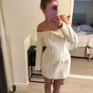 Vit offshoulder klänning - Snygg vit offshoulder klänning med knappar framtill. Klänningen har en figurnära passform och långa ärmar, perfekt för en stilren look. Passar utmärkt till student eller andra festliga tillfällen.