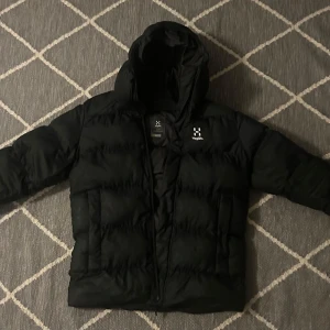 Svart dunjacka från Haglöfs - Säljer en svart dunjacka från Haglöfs med huva och dragkedja. Jackan är perfekt för kalla dagar och har en stilren design med långärmade ärmar. Den är vadderad för extra värme och komfort.