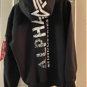 Svart hoodie från Alpha Industries - Säljer en svart hoodie från Alpha Industries med stor logga på ryggen och en röd detalj på ärmen. Tröjan har en klassisk känguruficka och ribbade muddar. Perfekt för en casual look.