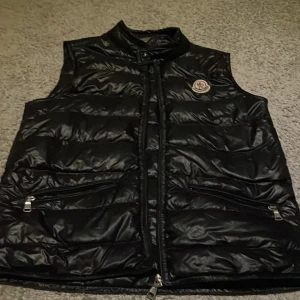 Svart dunväst från Moncler - Snygg svart dunväst från Moncler med dragkedja och två fickor framtill. Västen har en quiltad design och ett Moncler-märke på bröstet. Perfekt för kyligare dagar när du vill hålla stilen.