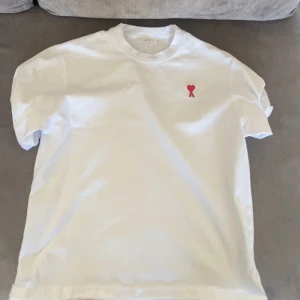 Vit t-shirt med rött hjärta - Säljer en stilren vit t-shirt med ett litet rött hjärta broderat på bröstet. Perfekt för en enkel och snygg look. T-shirten har korta ärmar och en klassisk passform.