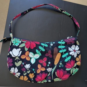 Blommig axelväska med dragkedja - Säljer en färgglad axelväska med blommigt mönster. Väskan har en dragkedja upptill och ett svart innerfoder. Perfekt för att liva upp din outfit med lite färg och mönster!