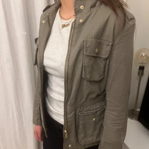 Grön parkajacka med dragkedja - Snygg grön parkajacka med dragkedja och flera praktiska fickor. Jackan har långa ärmar och en stilren design med knappar i guldton. Perfekt för vår och höst.