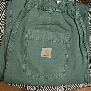  Carhartt judd pant - Säljer ett par gröna byxor från Carhartt med elastisk midja och en praktisk bakficka med logotyp. Perfekta för en avslappnad stil.