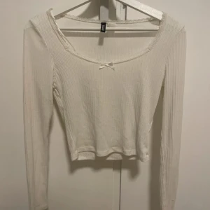 Vit ribbad topp från H&M Divided - Säljer en söt vit ribbad topp från H&M Divided. Toppen har långa ärmar och är dekorerad med spetsdetaljer och en liten rosett framtill.  Den är lite croppad i modellen 