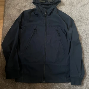 CP company jacka ny pris 4799kr - Denna jacka är en svart softshelljacka med en unik design. Den har en huva med inbyggda glasögon, vilket ger den en distinkt och futuristisk look. Jackan är försedd med en dragkedja framtill samt två diagonala fickor med dragkedjor på bröstet. Ärmarna har elastiska muddar, och materialet verkar vara både vatten- och vindavvisande, vilket gör den praktisk för olika väderförhållanden. Denna typ av jacka är ofta associerad med det populära märket C.P. Company