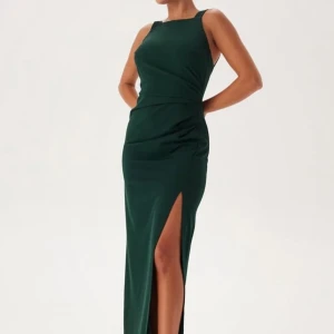 Mörkgrön långklänning med slits - Elegant mörkgrön långklänning med en stilren slits på sidan. Klänningen är ärmlös och har en figurnära passform som framhäver silhuetten. Perfekt för festliga tillfällen.