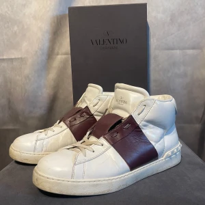 Höga Valentino Opens  - Snygga Valentino Garavani sneakers i vitt och vinrött. Skorna har en unik design med breda vinröda remmar över vristen och klassisk snörning. Perfekta för att ge din outfit en lyxig touch. Inget og  tillkommer. 