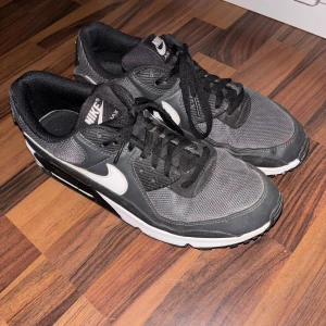 Nike Air Max 90 - Säljer ett par svarta Nike Air sneakers med vit sula och klassisk snörning. Skorna har en sportig design med Nike-loggan i vitt på sidan och tungan. Perfekta för en casual look.