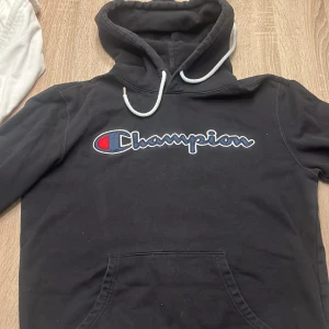 Svart hoodie från Champion - Säljer en svart hoodie från Champion med broderad logga på bröstet. Den har en klassisk känguruficka och justerbar huva med snören. Perfekt för en avslappnad stil. 