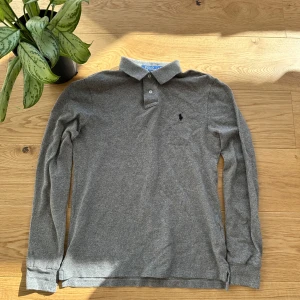 Grå pikétröja från Polo Ralph Lauren - Säljer en stilren grå pikétröja från Polo Ralph Lauren i slim fit. Tröjan har långa ärmar och en klassisk krage med knappar. Den ikoniska logotypen är broderad på bröstet. Perfekt för en avslappnad men ändå elegant look.