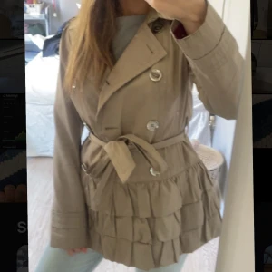 Beige trenchcoat med volangdetaljer - Snygg beige trenchcoat med knappar och bälte i midjan. Jackan har volangdetaljer nedtill som ger en unik look. Perfekt för vår och höst!