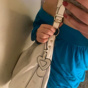 Vit axelväska med blommigt foder - Snygg vit axelväska i syntetmaterial med justerbara remmar och dekorativt hjärtdetalj. Väskan har ett blommigt foder och flera praktiska fack med dragkedja för enkel organisering.