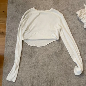 Vit topp från NA-KD - Säljer en stilren vit topp från NA-KD. Toppen har långa ärmar och en bekväm passform som passar perfekt till jeans eller kjol. Perfekt för en trendig look! Men toppen är också väldigt kort