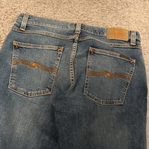 Blå jeans från Nudie Jeans - Snygga blå jeans från Nudie Jeans med klassisk femficksdesign och orange sömmar. De har en rak passform och är perfekta för en avslappnad stil. Märkeslogga på baksidan och knappgylf framtill.