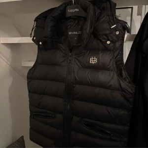 Byte söks  - Snygg svart dunväst från KappAhl med dragkedja och två fickor framtill. Västen har en hög krage och en broderad logotyp på bröstet. Perfekt för kyligare dagar.