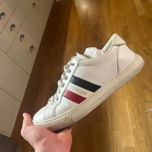 Vita sneakers från Moncler - OBS köp inte skriv till mig innan!!! Snygga vita sneakers från Moncler med klassiska ränder i svart och rött på sidan. Skorna har snörning och en stilren design som passar perfekt för en trendig look. Ny pris runt 6 tusen!!