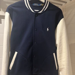 Blå och vit jacka från Polo Ralph Lauren - Säljer en snygg blå och vit jacka från Polo Ralph Lauren. Jackan har långa vita ärmar och en blå kropp med vita detaljer. Den har knappar framtill och ribbade muddar vid ärmslut och nederkant. Strl: M, säljes pga. rensar garderoben, aldrig använd!