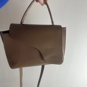 Brun handväska från ATP Atelier - Snygg brun handväska från ATP Atelier i skinn. Väskan har ett elegant lock och ett praktiskt handtag för enkel bärning. Perfekt för att ge din outfit en stilren touch. Är i den storka storleken, ny pris 5200