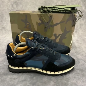 Valentino Rockrunner sneakers i camouflage - Snygga Valentino Rockrunner sneakers i en cool camouflage-design. Skorna har en mörkblå och svart färgkombination med ikoniska nitar runt sulan. Tillverkade i en mix av mocka och syntetmaterial, dessa sneakers har snörning och en bekväm passform. Obs! (ENDAST SKORNA TILLKOMMER) Vid frågor och funderingar, tveka inte på att höra av er!
