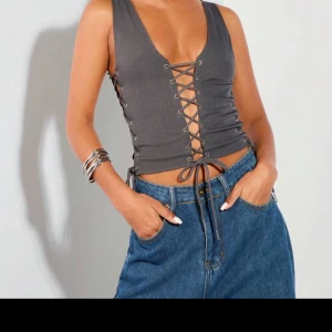 Grå korsettväst med snörning - Snygg grå korsettväst med snörning på sidorna och fram. Perfekt för att ge en edgy touch till din outfit. Västen har en v-ringad design och är tillverkad i ett stilrent material.