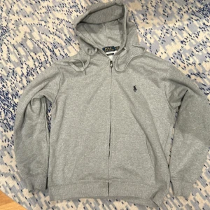 Grå hoodie från Polo Ralph Lauren - Säljer en stilren grå hoodie från Polo Ralph Lauren med dragkedja och det klassiska logotypbroderiet på bröstet. Tröjan har en justerbar huva och ribbade muddar för extra komfort. Perfekt för en avslappnad look💯. Tröjan är i väldigt bra skick och jag bedömer att den är true to size.📐Om du har några frågor är det bara att skicka ett meddelande så svarar jag så fort som möjligt😊👍