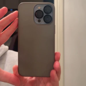 Mobilskal till iPhone - Säljer ett stilrent grått mobilskal som passar till iPhone 13 PRO. Skalet är gjort i mjuk syntet och har en matt finish. Det täcker baksidan och kanterna på mobilen och har ett stort utskuret hål för kameran. Perfekt för dig som vill ha ett diskret och skyddande skal.
