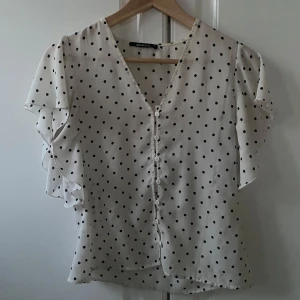 Vit blus med svarta prickar och volangärmar - Säljer min jättefina vita blus med svarta prickar, som är ifrån Ginatricot. Blusen är ca 2 år gammal, men är i perfekt skick! Varför jag säljer denna blusen är för att jag inte har någon användning för den, och eftersom prickigt mönster har blivit populärt igen passar jag på att sälja den!🥰