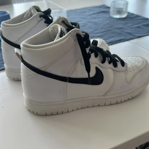 Nike Dunk High vita och svarta sneakers - Säljer ett par klassiska Nike Dunk High sneakers i vitt med svarta detaljer och svart swoosh. Skorna har högt skaft, svart snörning och rund tå. Perfekta för dig som gillar streetwear och vill ha en stilren look.